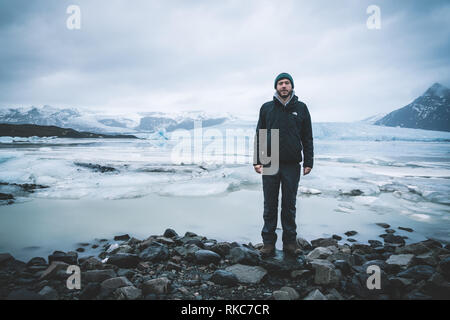 Giovane uomo in piedi di fronte a Fjallsarlon laguna iceberg, all'estremità sud del ghiacciaio Vatnajokull, con iceberg galleggianti che calve) dal bordo Foto Stock