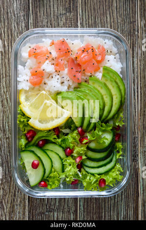 Scatola di pranzo: riso, salmone, con insalata di cetrioli, avocado, verdi, limone, chia semi su un sfondo di legno. Cibo per il fitness. Il concetto di mangiare sano. Foto Stock