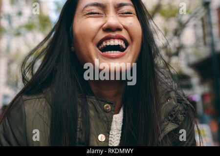 Ritratto di una donna che ride con gli occhi chiusi. Tagliate il colpo di una donna asiatica di ridere in piedi all'esterno. Foto Stock