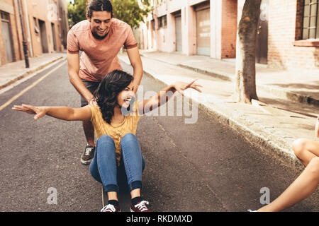 Excited donna giovane essendo spinta su skateboard con il suo fidanzato all'aperto sulla strada, con gli amici seduti lungo la strada. Giovane che si diverte ou Foto Stock