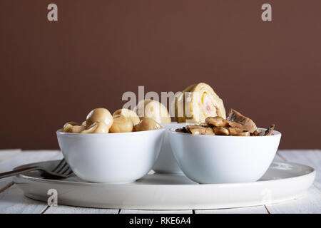 I funghi e i carciofi in olio nella coppa in ceramica bianca su sfondo di legno. Foto Stock