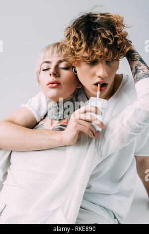 La donna tatuata abbracciando uomo bello con capelli ricci azienda sigaretta in bocca isolato su grigio Foto Stock