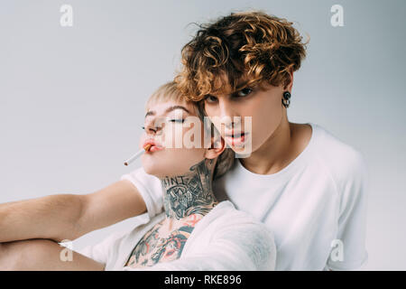 La donna tatuata holding sigaretta in bocca vicino all uomo bello con capelli ricci isolato su grigio Foto Stock