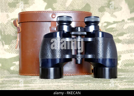 Vintage militare inglese a prisma di Porro colore nero binocolo con aperto brown una custodia in pelle con tracolla su sfondo di mimetizzazione vista frontale vicino Foto Stock