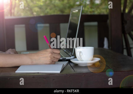 Una donna scrivere nota sul notebook e laptop aperto con tazza di caffè sul tavolo di legno. Foto Stock
