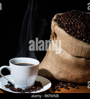 Macchine da caffè in una tazza bianca con vapore o vapore e i chicchi di caffè in una tela di sacco di Hesse e fagioli sparsi su una superficie di legno. Foto Stock