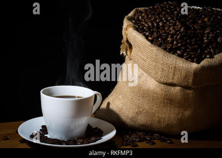 Macchine da caffè in una tazza bianca con vapore o vapore e i chicchi di caffè in una tela di sacco di Hesse e fagioli sparsi su una superficie di legno. Foto Stock