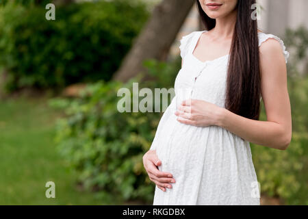 Ritagliato shot di sorridere donna incinta in abito bianco in piedi in posizione di parcheggio Foto Stock