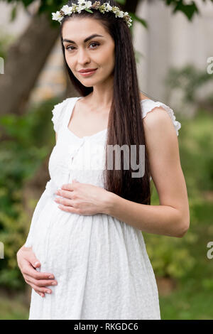 Bella felice giovane donna incinta in abito bianco e ghirlanda floreale sorridente in telecamera in posizione di parcheggio Foto Stock