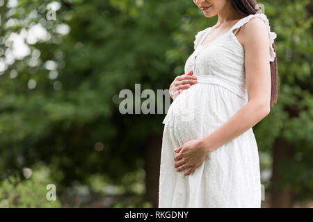 Ritagliato colpo di gravidanza sorridente giovane donna in abito bianco toccando ventre in posizione di parcheggio Foto Stock