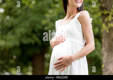 Ritagliato shot di sorridere donna incinta in abito bianco toccando ventre in posizione di parcheggio Foto Stock