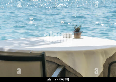 Tabella vuota in un cafè sulla riva del mare o del lago, coperto con una tovaglia bianca con un posacenere e un fiore decorativo sulla pentola una soleggiata giornata estiva. Foto Stock