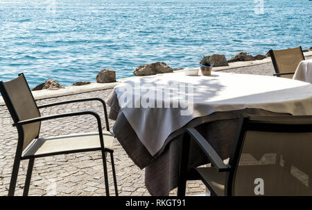 Tabella vuota in un bar in riva al mare o al lago, coperto con una tovaglia bianca con posacenere e un fiore decorativo sulla pentola una soleggiata giornata estiva. Foto Stock