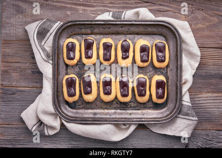 Mini eclairs alla vaniglia con glassa di cioccolato sul vassoio metallico, vista dall'alto in legno rustico Foto Stock