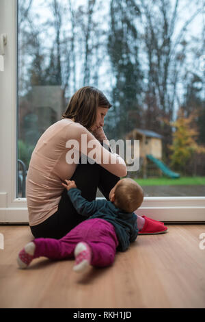 Giovane madre seduta preoccupato o premuto mediante la finestra in una stanza vivente con la sua bimba sdraiato sul pavimento raggiungendo fuori a lei. Foto Stock