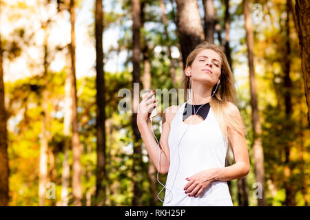 Bella bionda modello in abbigliamento da palestra ascolta musica sul suo telefono cellulare. Sfocata sullo sfondo di foresta al tramonto Foto Stock