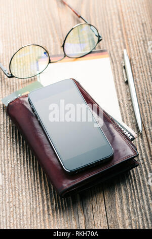 Smartphone con schermo vuoto, portafogli, dollaro banconote, carte di credito e carte di notebook su un tavolo di legno. Vista da sopra. Orientamento verticale Foto Stock