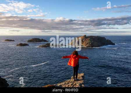 Woman in Red jacket è in piedi sul bordo di una scogliera con le braccia aperte e godere degli splendidi paesaggi dell'oceano. Preso in testa di corvo, Nord Twillingate Foto Stock