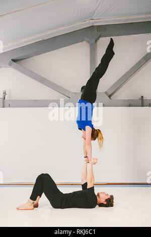 Il tema è lo sport e gli acri. Un giovane caucasico maschio e femmina giovane praticante yoga acrobatico in una palestra bianca su stuoie. Un uomo giace sulla sua schiena e hol Foto Stock