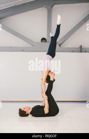 Il tema è lo sport e gli acri. Un giovane caucasico maschio e femmina giovane praticante yoga acrobatico in una palestra bianca su stuoie. Un uomo giace sulla sua schiena e hol Foto Stock
