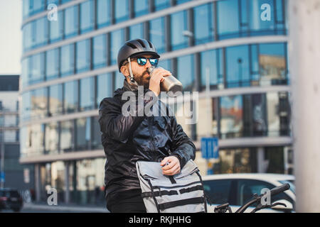 Corriere maschio con occhiali da sole di bere acqua quando la consegna di pacchi in città. Foto Stock