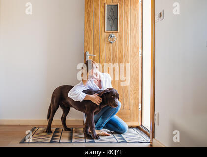 Una giovane donna seduta al chiuso dalla porta sul pavimento a casa, giocando con un cane. Foto Stock