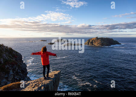 Woman in Red jacket è in piedi sul bordo di una scogliera con le braccia aperte e godere degli splendidi paesaggi dell'oceano. Preso in testa di corvo, Nord Twillingate Foto Stock