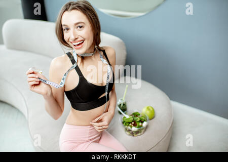 Ritratto di una bella donna sportive con cibo sano e nastro di misurazione sul divano di casa. Uno stile di vita sano e perdita di peso concept Foto Stock