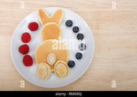 Frittelle in un coniglietto coniglio pattern, banana per la coda e i piedi, fresche lamponi e mirtilli sulla piastra su un legno chiaro Tavolo con spazio di copia Foto Stock