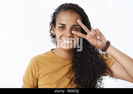 Close-up shot di successo e piacere piuttosto giovani afro-american donna gentile con scuri lunghi capelli ricci che mostra la pace o segno di vittoria su Foto Stock