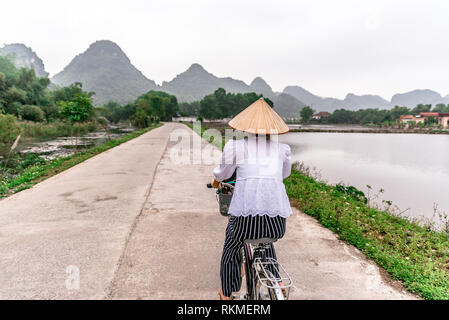 Donna vietnamita con il tradizionale conica di cappello di paglia sulla bicicletta. Lo splendido paesaggio di campi di riso e del paesaggio di montagna nella riserva naturale di Trang Un e Foto Stock