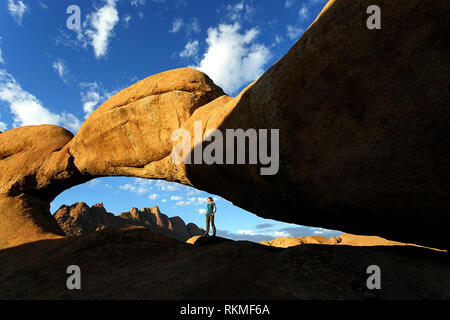 Donna in piedi sotto la roccia naturale arch vicino alla montagna di granito Spitzkoppe con ottima vista sulle montagne sullo sfondo, Namibia, Africa Foto Stock