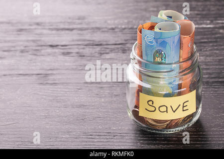 Le banconote e le monete in euro in un vasetto di vetro su un tavoli in legno nero lucido con la riflessione su una superficie lucida. Risparmiare denaro concept Foto Stock