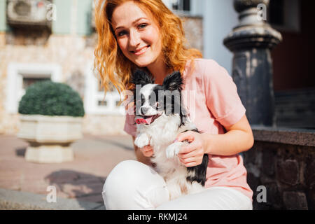 Il tema è l amicizia di uomo e animali. Giovane e bella con i capelli rossi donna caucasica tenendo un cane di razza Chihuahua in prossimità di una costruzione casa in Foto Stock