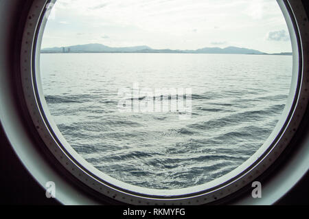 Oblò rotondo su una nave da crociera, vista interna attraverso la finestra sulla costa e sul mare, sunrise contro il mare, close-up. Foto Stock