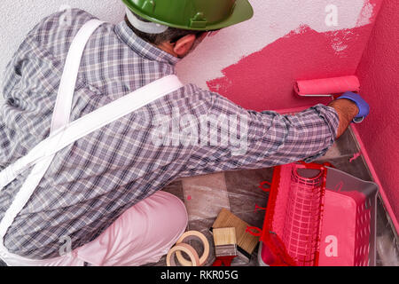 Casa caucasica pittore lavoratore in bianco tute da lavoro, con il rullo che dipinge la parete con la pittura colorata di rosa. Industria edile. A Foto Stock