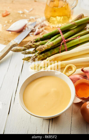 Una deliziosa tazza di salsa olandese con grappoli di fresco verde e asparagi bianchi su un tavolo da cucina con le uova incrinate con i tuorli per la salsa Foto Stock