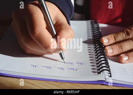 Giovane donna seduti ad una scrivania, la scrittura di un elenco di propositi per l'anno nuovo in una rilegatura a spirale notebook, a partire da gennaio a secco. Sezione mediana, close-up mano Foto Stock
