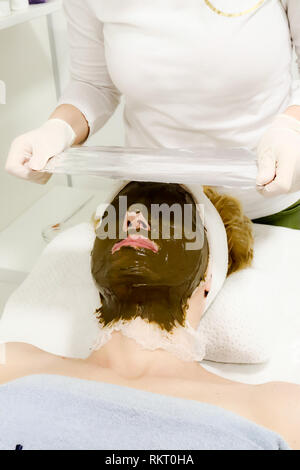 Estetista lavoratore mette un foglio di alluminio sul cliente femmina faccia, procedura dopo l'applicazione di cosmetici per il viso maschera di mare ​​algae Foto Stock
