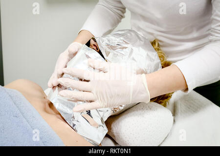 Estetista lavoratore mette un foglio di alluminio sul cliente femmina faccia, procedura dopo l'applicazione di cosmetici per il viso maschera di mare ​​algae Foto Stock