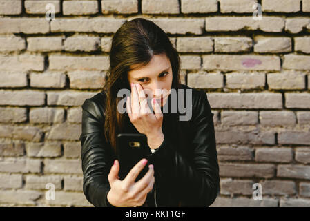 Giovane ragazza utilizzando lo schermo del suo cellulare come uno specchio per mettere il trucco e rimuovere brufoli dal suo volto e lei ottiene abbastanza per una data con il suo Foto Stock