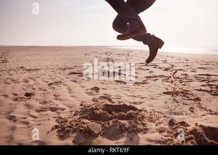 Uomo che indossa jeans e scarpe con tacco, saltare su di una spiaggia sabbiosa spiaggia del Regno Unito in una fredda giornata di primavera Foto Stock