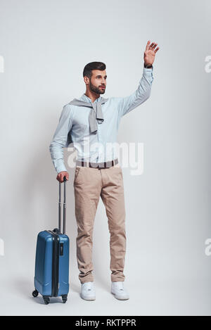 Taxi. Giovane e bello uomo con un bagaglio è alzando la mano mentre in piedi in studio su sfondo grigio. Lunghezza completa foto. Concetto di viaggio Foto Stock