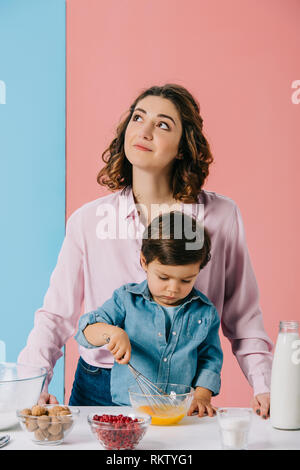 Madre cercando e sorridere mentre piccolo figlio di uova di mantecazione su sfondo bicolore Foto Stock