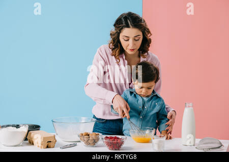 Bella madre con carino piccolo figlio di monta le uova in una terrina insieme sullo sfondo bicolore Foto Stock