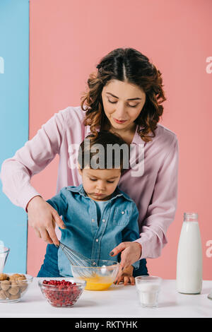 Bella madre aiutare carino piccolo figlio in uova di mantecazione con palloncino frusta in vaso su sfondo bicolore Foto Stock