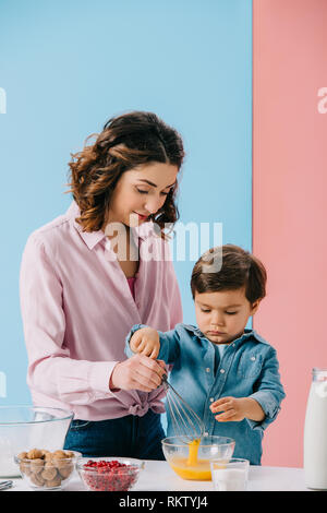 Madre aiutare piccolo figlio in uova di mantecazione con palloncino frusta su sfondo bicolore Foto Stock