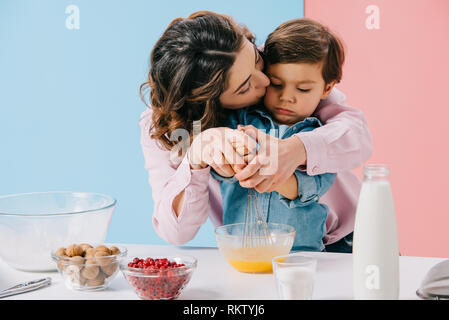 Mommy kissing adorabili poco figlio mentre le uova di mantecazione con palloncino frusta insieme sullo sfondo bicolore Foto Stock