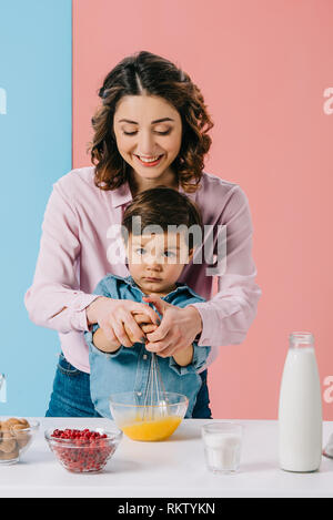 Madre sorridente con carino piccolo figlio di uova di mantecazione con palloncino frusta durante la cottura insieme sullo sfondo bicolore Foto Stock