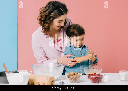 Sorridente madre aiutare piccolo figlio in uova di mantecazione con palloncino frusta su sfondo bicolore Foto Stock